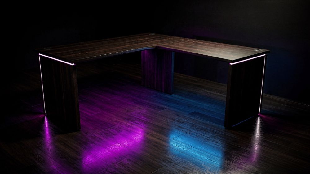 Mesa gamer em L de madeira escura com iluminação RGB roxa e azul refletindo no chão, estilo Nerdesk